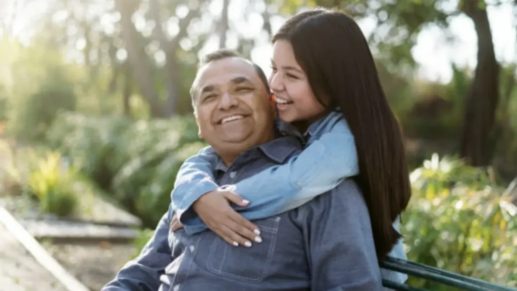 A young girl smiles while hugging a man from behind, both enjoying an outdoor moment amidst trees and greenery at Chattleton Place.
